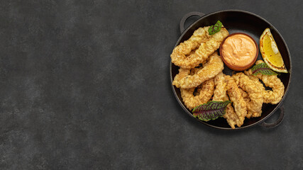 Golden rings of squid fries in a round frying pan with sauce, herbs and lemon on a dark background...