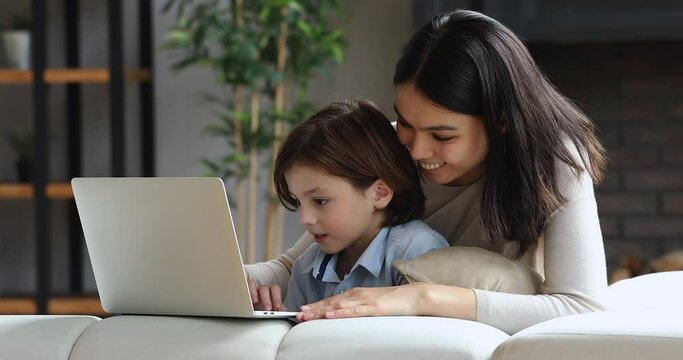 Lovely young Asian babysitter teach little boy to use laptop. Multi ethnic family relax on sofa at home enjoy online fun, choose goods buy on e-commerce services. Modern tech, kids development concept