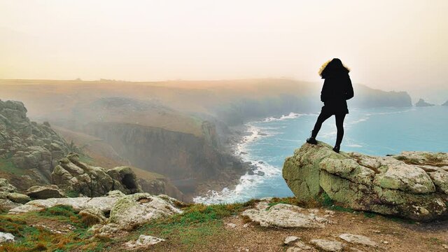 Young woman standing on the edge of a rocky cliff and looking at the blue ocean. A girl standing and enjoying a scenic beautiful crystal clear sea water in Cornwall, England in 4K.