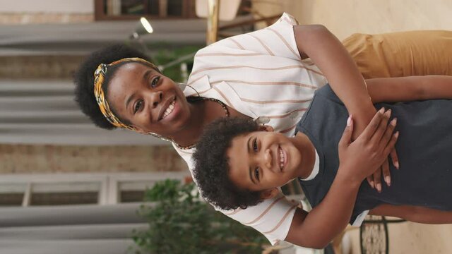 Vertical medium portrait with slowmo of caring afro-american woman hugging cute 8-year-old son smiling to camera standing in cozy living room