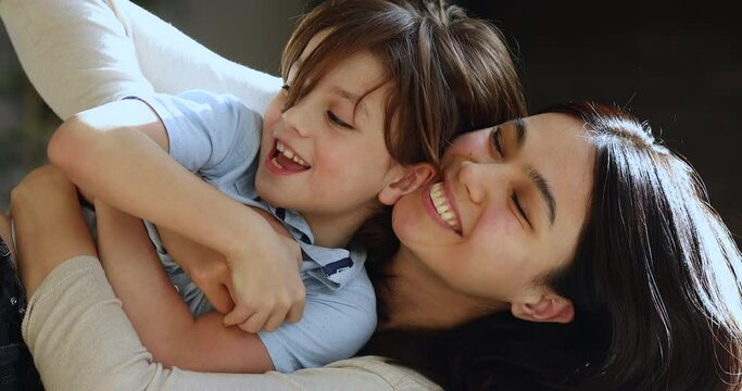 Close up Asian woman cuddling tickling little son, multi ethnic family lying on couch enjoy priceless time on weekend together. Adoption and foster, new loving mom for adorable preschool child concept
