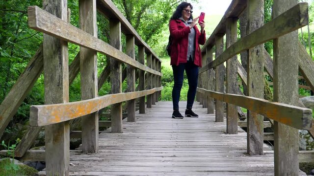 City woman with eyeglasses and long hair wearing red jacket walking on small wooden footbridge towards camera, stops in the middle to take some pictures.