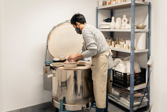 Woman potter with mask putting pottery in kiln.