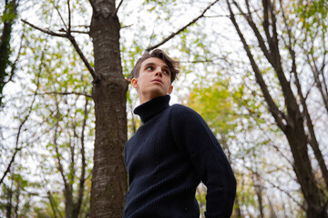 A man stands in an autumn forest, wearing a black wool sweater with a high collar and looking around.Thoughtful young male in a wood.