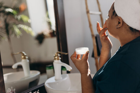 Black woman applying cream on face in bathroom