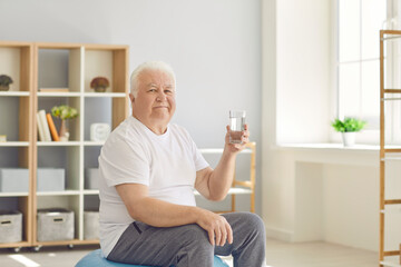 Senior man holding a glass of water sitting on a fitball at home and looking at the camera. Concept of active lifestyle and maintaining water balance in the elderly.