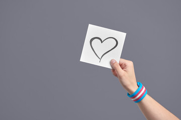 Young hand holding a paper with a heart, wearing a transgender flag bracelet