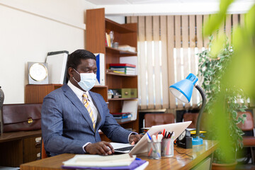 African american businessman wearing protective face mask focused on working with laptop in modern office. Necessary precautions during coronavirus pandemic