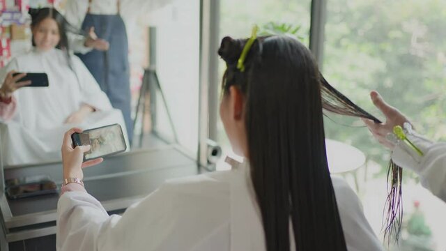 Asian woman use phone take photo while hairdresser cut hair in salon.
