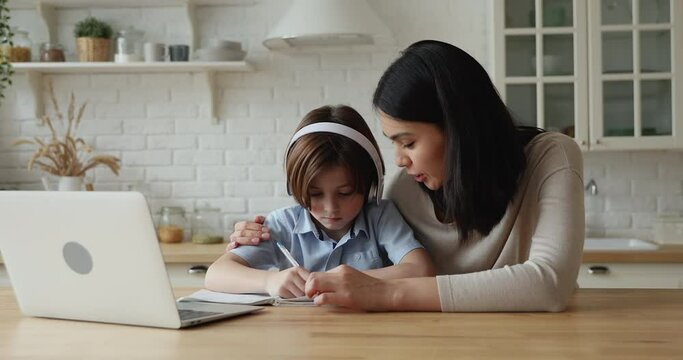 7s boy wear headphones take notes in copybook doing homework receiving guidance by young Asian female tutor. Mom helps to son with hometask primary school assignment praising giving high five concept