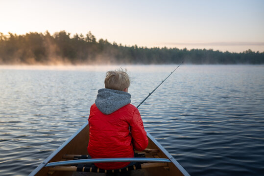 Early morning Muskoka lake mist rises as boy fishes ffrom canoe