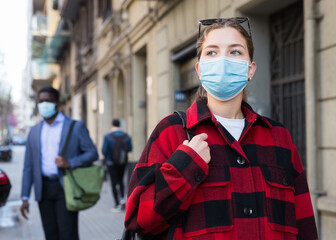 Young woman in disposable face mask walking along city street. Concept of Covid 19 virus spread...