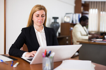 Focused young woman wearing business suit sitting at desk in modern office interior, working with laptop