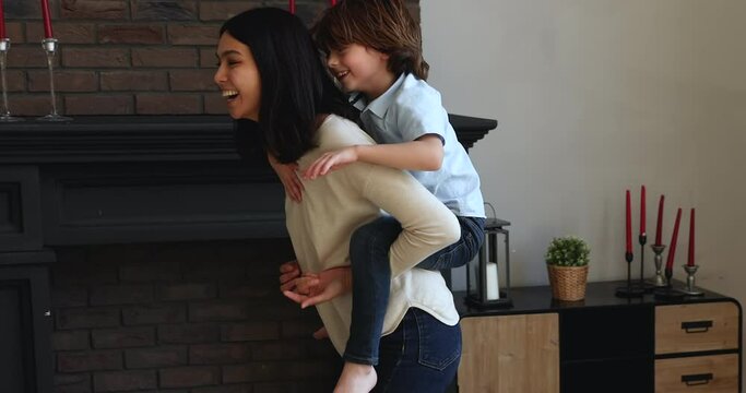 Happy family young asian mom play piggyback cute little 6s funny adopted son, nanny carry boy on back run in cozy living room enjoy playtime spend active weekend together at home. Fun, leisure concept