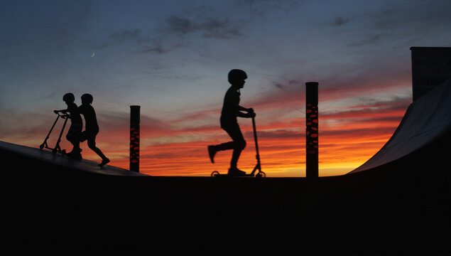 Boy riding kick scooter on ramp near friends at sunset