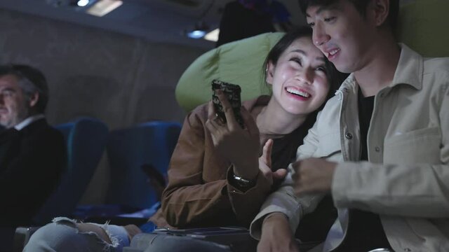 Young Asian couple using smartphone together happily while seating in business class during traveling by plane at night. Transportation or technology lifestyle concept.