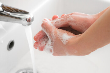Fototapeta premium Hands of woman wash their hands in a sink with foam to wash the skin and water flows through the hands. Concept of health, cleaning and preventing germs and coronavirus from contacting hands