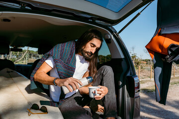 man with hot beverage sitting on car trunk