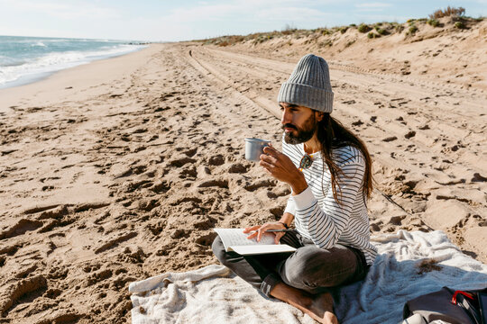 long hair men sitting on beach coast writing on notebook and drinking coffee