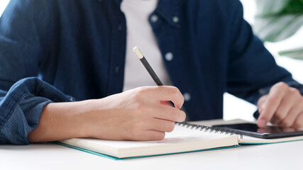 Fototapeta premium Online study, Man hand writing on notebook while using mobile phone for digital home study class, Adult male student doing lesson while e learning by smartphone at house, Education