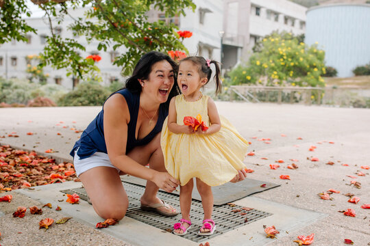 Mom laughs as air billows yellow dress of young Asian girl holding orange flowers