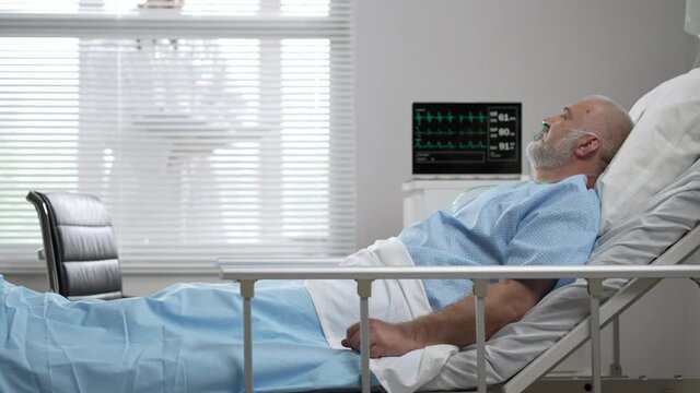 In the clinic, an elderly man, the patient is lying on the bed connected to the ECG machine and the doctor is standing outside the window of the ward and looks at the indicators. 