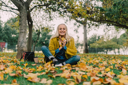 Senior woman sitting on the park floor holding dry leaves and looking at camera