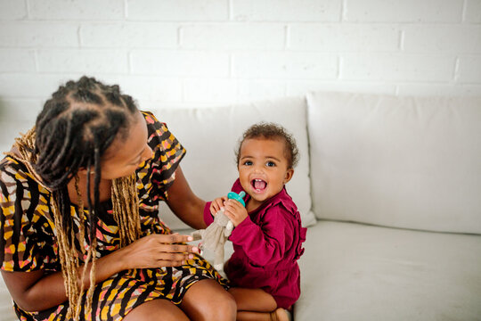 Stylish black mom with long braids sitting on white couch next to happy biracial