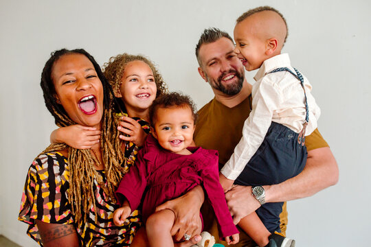 Laughing biracial family with three young children held by dad and mom indoors with neutral background.