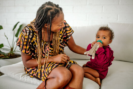 Trendy black mom with dreadlocks sitting on white couch next to 1 yr old biracial baby 
