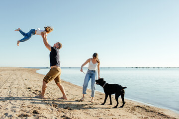 Happy young family with dog resting on sandy seashore against cloudless blue sky