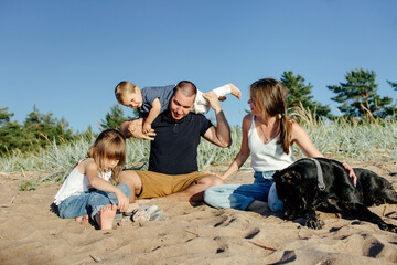Cute joyful children resting on sandy beach with young parents and obedient dog