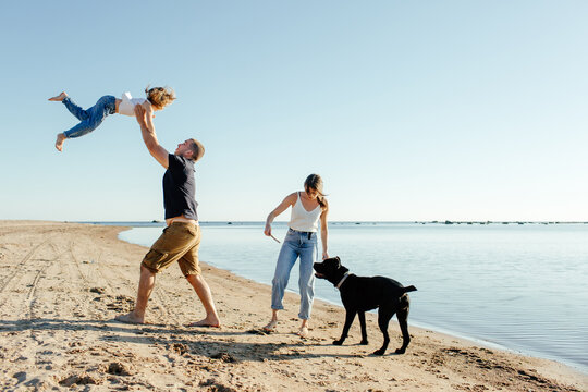 Happy young family with dog resting on sandy seashore against cloudless blue sky