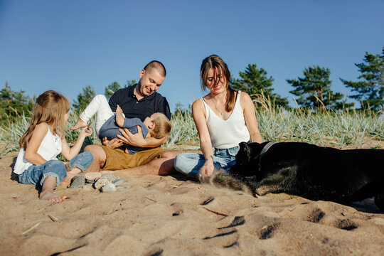 Cute Joyful Children Resting On Sandy Beach With Young Parents And Obedient Dog