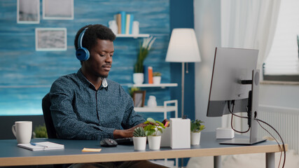 Obraz na płótnie Canvas African american black manager using headphones to listen music while working from home office on computer PC. Young freelancer working man computer user listening and enjoying music in home office