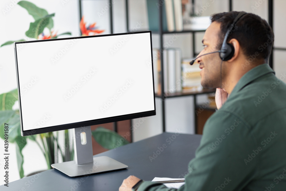 Wall mural happy indian businessman in headset looking at blank computer screen, studying at home concept dista