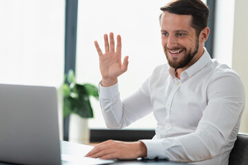 Confident successful young caucasian businessman using laptop to meeting online with colleagues or partners in the office, video call, smiling, reeting gesture