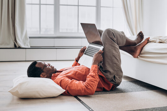 Barefoot African-American man freelancer in orange shirt works on modern laptop lying on floor near comfortable bed side view