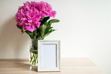 Portrait white picture frame mockup on wooden table. Modern vase with peony. White wall background. Scandinavian interior. 