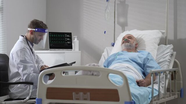 Young doctor talking to senior man resting in hospital bed. Male RN assisting elderly male patient lying down in bed. Friendly male Doctor Checks on the Sick Male Patient Lying in Bed