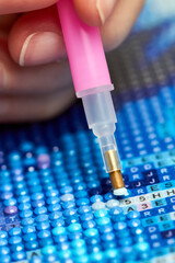 Girl collect diamond painting. Diamond embroidery with a tweezers. Acrylic rhinestones. Close-up, selective focus.