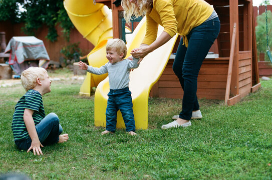 Mom helps happy young son walk to big brother near backyard playset 