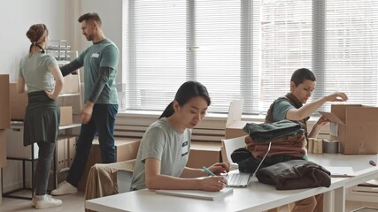 Medium slowmo of small group of people volunteering in office – packing food and clothes for homeless or needy people into boxes and chatting with each other - Powered by Adobe