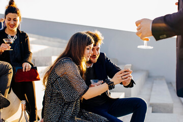 Interested well dressed colleagues sharing smartphone during party on stairs