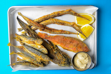 Fried fish with sauce and lemon served on a metal tray over blue background.
