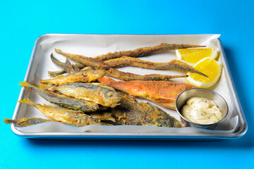 Fried fish with sauce and lemon served on a metal tray over blue background.