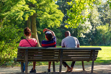 Fototapeta premium Three people sitting on a bench in a sunny and green Amsterdam Vondelpark in the Netherlands.