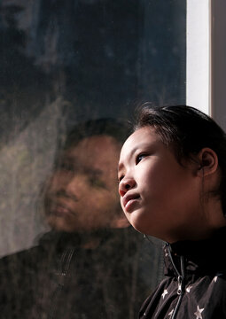 Lovely little asian girl, by the window at sunset. The window glass reflected herself