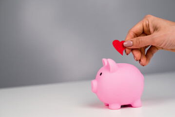 Faceless caucasian woman folds a small heart into a pink piggy bank on a white background. Love...