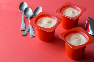 Red colour yogurt plastic pots with spoons and foil on red background - creamy fruit yoghurt in plastic cups with copy space